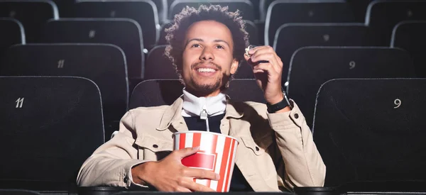 Plan panoramique d'un homme afro-américain souriant tenant du pop-corn et regardant un film au cinéma — Photo de stock