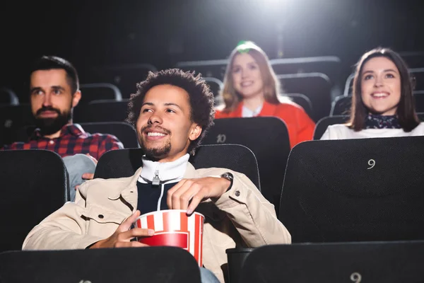 Foco seletivo de sorrir afro-americano segurando pipocas e assistindo filme no cinema — Fotografia de Stock