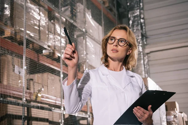 Attractive storekeeper holding clipboard, looking away and pointing with pen in warehouse — Stock Photo