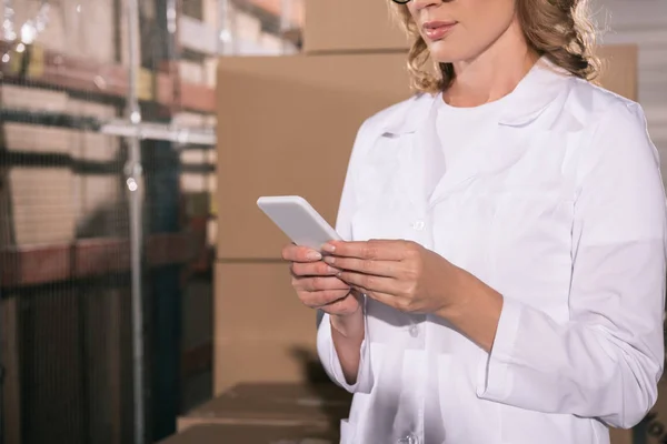 Cropped view of storekeeper in white coat using smartphone in warehouse — Stock Photo