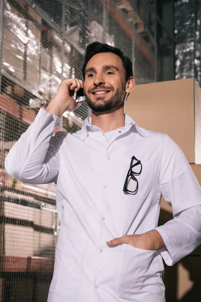 Lächelnder Ladenbesitzer, der mit dem Smartphone spricht, während er mit der Hand in der Tasche im Lager steht — Stockfoto