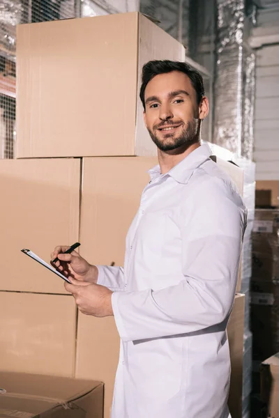 Handsome storekeeper in white coat smiling at camera while writing on clipboard in warehouse — Stock Photo