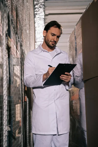 Focused storekeeper in white coat writing on clipboard in warehouse — Stock Photo