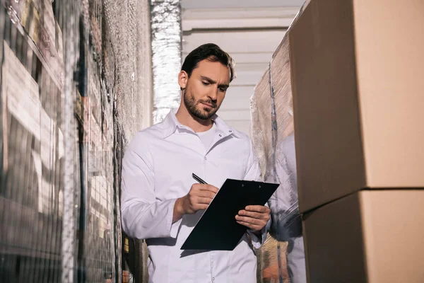 Concentrated storekeeper in white coat writing on clipboard in warehouse — Stock Photo