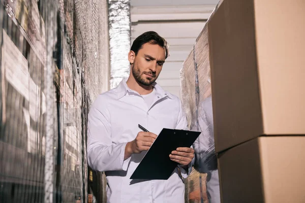 Konzentrierter Lagerist in weißer Garderobe mit Schrift auf Klemmbrett im Lager — Stockfoto