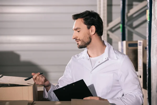 Handsome storekeeper inspecting cardboard boxes while holding clipboard and pen — Stock Photo