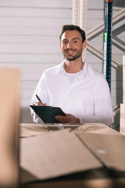 Foco seletivo do armazenista sorrindo segurando prancheta enquanto estiver perto de caixas de papelão — Fotografia de Stock