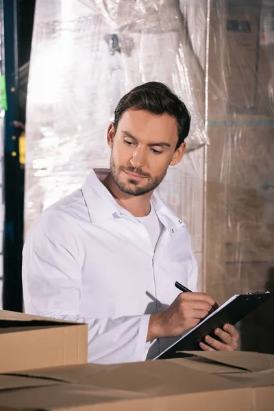 Concentrated storekeeper writing on clipboard while looking at packages in warehouse — Stock Photo