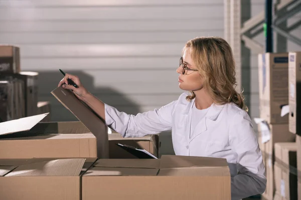 Focused storekeeper in white coat inspecting cardboard boxes in warehouse — Stock Photo