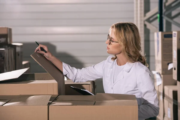 Gardien concentré en manteau blanc inspectant les boîtes en carton dans l'entrepôt — Photo de stock