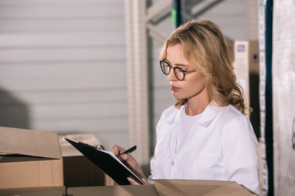 Concentrated storekeeper in glasses writing on clipboard in warehouse — Stock Photo