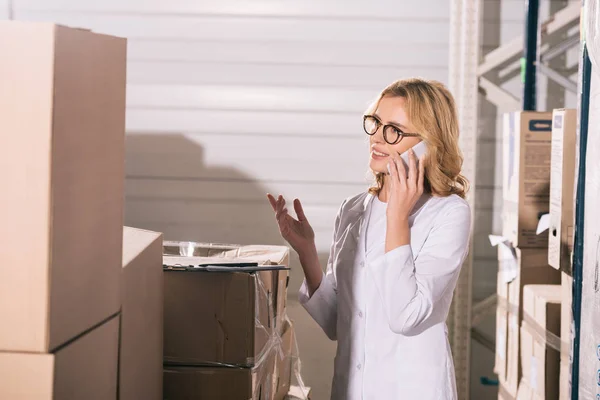 Smiling storekeeper talking on smartphone and showing question gesture while looking at cardboard boxes in warehouse — Stock Photo