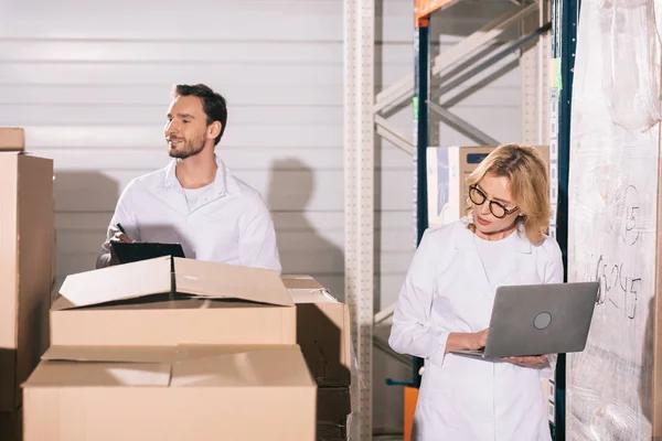 Attractive storekeeper using laptop near handsome colleague writing on clipboard in warehouse — Stock Photo