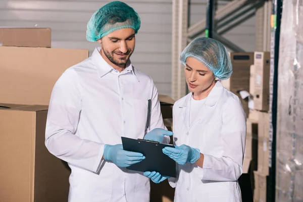 Two positive storekeepers in white coats and hairnets looking at clipboard in warehouse — Stock Photo