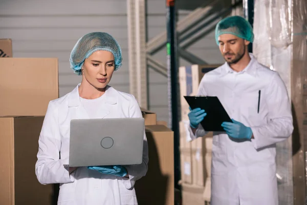 Armazenista atraente usando laptop perto colega olhando para a área de transferência no armazém — Fotografia de Stock