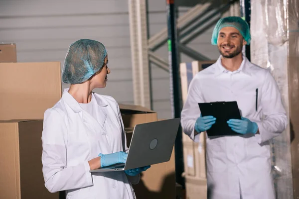 Sorrindo armazenista segurando prancheta e olhando para colega usando laptop no armazém — Fotografia de Stock