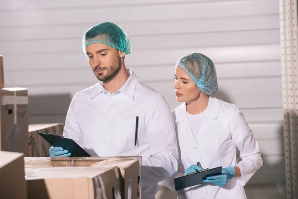 Atractivo tendero mirando portapapeles en manos de un colega guapo mientras está de pie cerca de cajas de cartón - foto de stock