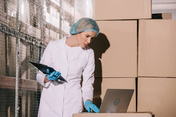 Concentrated storekeeper in white coat and hairnet using laptop and holding clipboard — Stock Photo