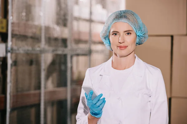 Attractive storekeeper in hairnet smiling at camera while holding smartphone — Stock Photo