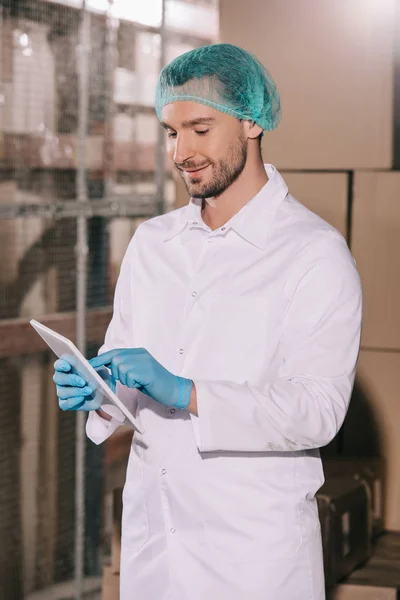 Magasinier souriant en manteau blanc et filet à cheveux en utilisant une tablette numérique dans l'entrepôt — Photo de stock