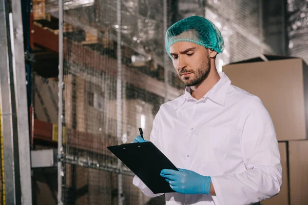 Concentrated storekeeper in white coat and hairnet writing on clipboard in warehouse — Stock Photo