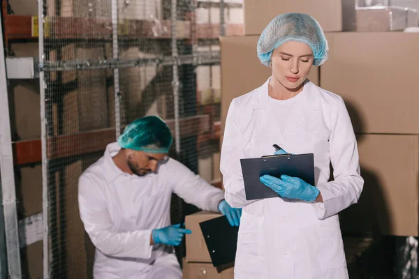 Attractive storekeeper writing on clipboard near colleague inspecting cardboard box — Stock Photo