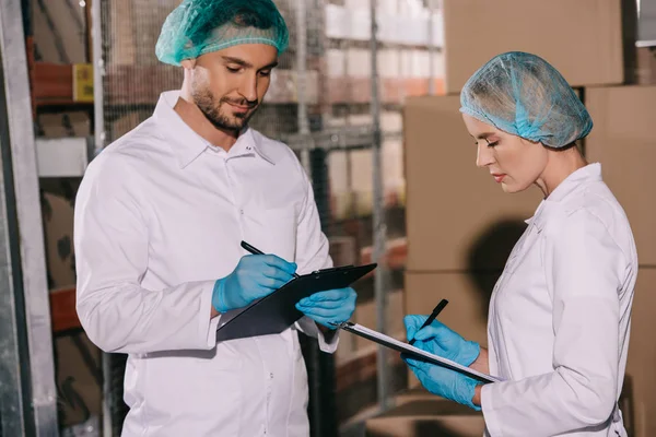 Two storekeepers in white coats and hairnets writing on clipboards in warehouse — Stock Photo
