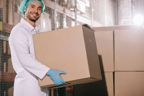 Cheerful storekeeper smiling at camera while holding cardboard box in warehouse — Stock Photo