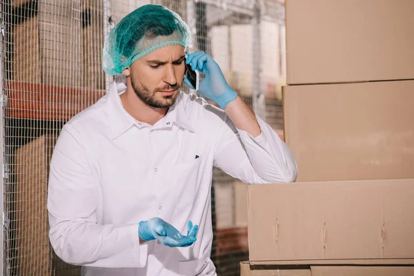 Almacenista serio hablando en el teléfono inteligente mientras está de pie cerca de cajas de cartón en el almacén - foto de stock