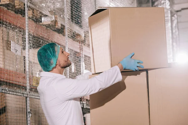 Storekeeper in white coat and hairnet taking cardboard box in warehouse — Stock Photo