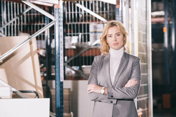 Confident businesswoman looking at camera while standing in warehouse with crossed arms — Stock Photo