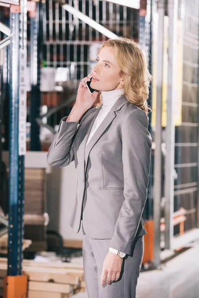 Serious businesswoman talking on smartphone and looking away in warehouse — Stock Photo