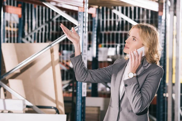 Femme d'affaires sérieuse parlant sur smartphone, détournant les yeux et pointant la main dans l'entrepôt — Photo de stock