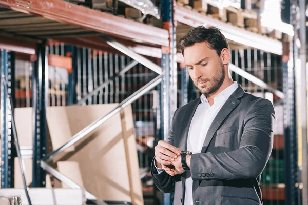 Hombre de negocios serio comprobar el tiempo en reloj de pulsera mientras está de pie en el almacén - foto de stock