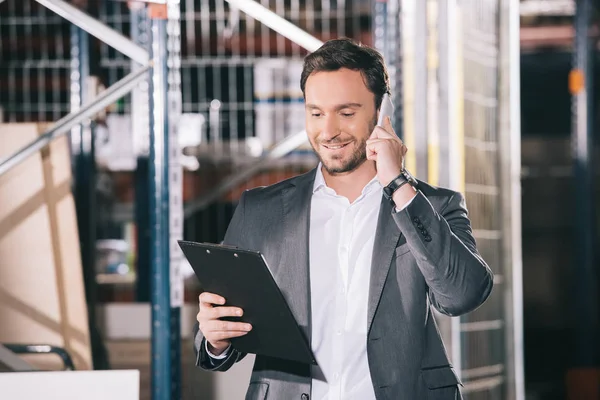 Smiling businessman talking on smartphone and looking on clipboard in warehouse — Stock Photo