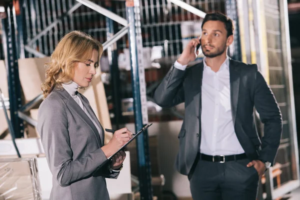 Empresaria concentrada escribiendo en portapapeles cerca de hombre de negocios hablando en el teléfono inteligente en el almacén — Stock Photo