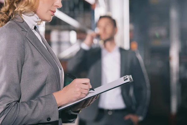 Selektiver Fokus der Geschäftsfrau, die auf Klemmbrett schreibt, in der Nähe eines Geschäftsmannes, der im Lager mit dem Smartphone spricht — Stockfoto