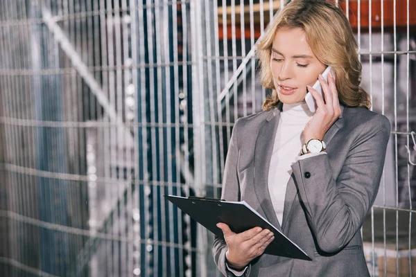 Atractiva mujer de negocios hablando en el teléfono inteligente y sujetando portapapeles en el almacén - foto de stock