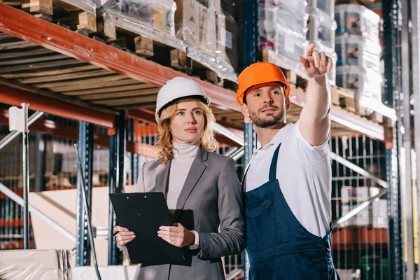 Guapo trabajador de almacén apuntando con la mano mientras está de pie cerca de la mujer de negocios en el almacén - foto de stock