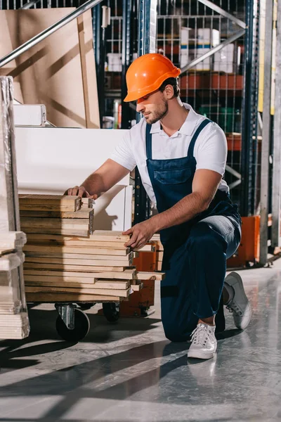 Ouvrier d'entrepôt debout sur le genou près du rack avec des planches en bois — Photo de stock