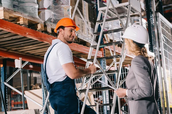 Sorrindo trabalhador armazém em pé na escada e olhando para mulher de negócios no capacete — Fotografia de Stock