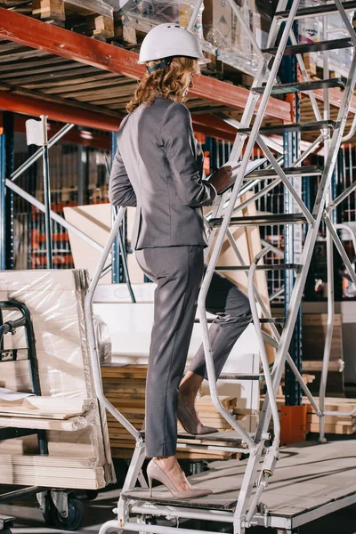 Geschäftsfrau in offizieller Kleidung und Helm steht auf Leiter in Lagerhalle — Stockfoto