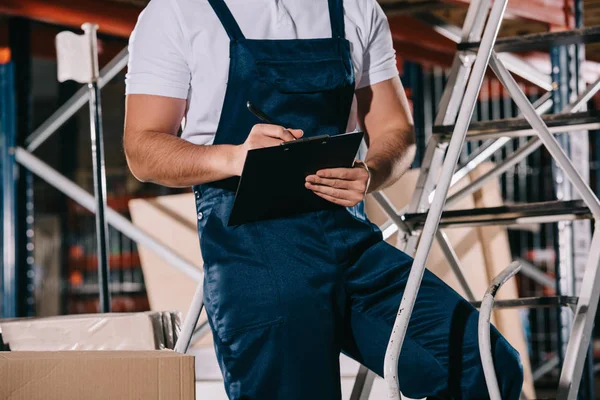 Cropped view of warehouse worker in overalls writing on clipboard in warehouse — Stock Photo