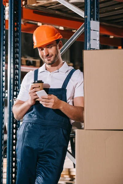 Smiling loader holding coffee to go and using smartphone in warehouse — Stock Photo