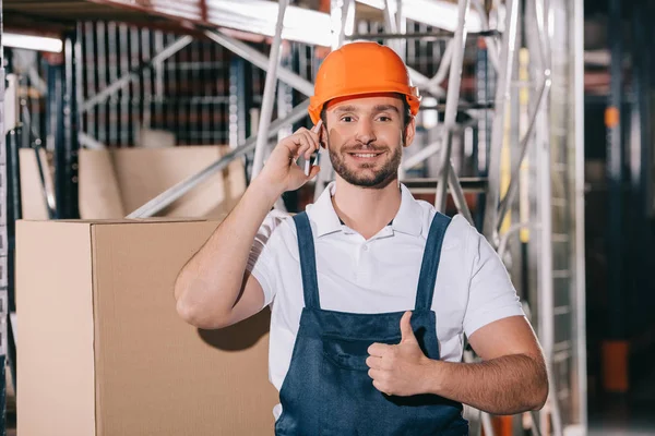 Sorrindo trabalhador armazém falando no smartphone, olhando para a câmera e mostrando o polegar para cima — Fotografia de Stock