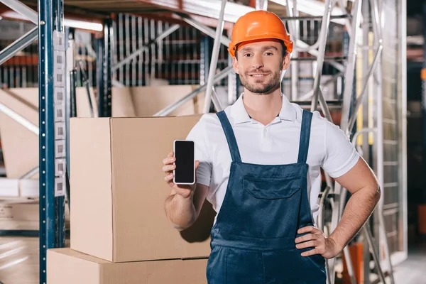 Chargeur souriant debout avec la main sur la hanche et montrant smartphone avec écran vide — Photo de stock