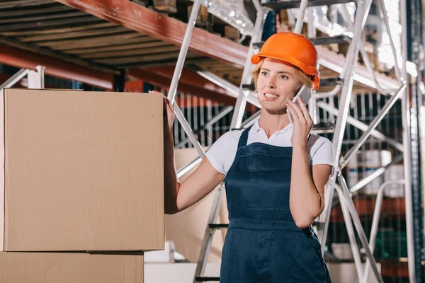 Trabajadora sonriente hablando en el teléfono inteligente mientras está de pie cerca de cajas de cartón - foto de stock