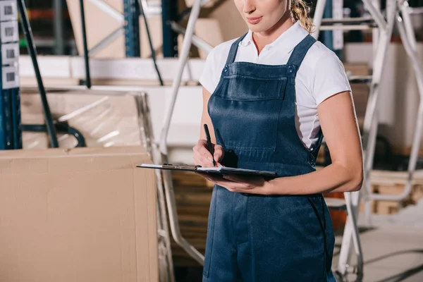 Vista ritagliata di operaia in tuta da lavoro scrivere sugli appunti in magazzino — Foto stock