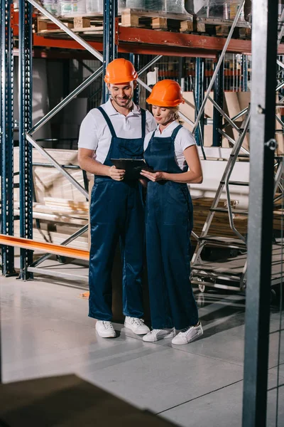 Travailleurs souriants entrepôt en salopettes et casques regardant sur presse-papiers ensemble — Photo de stock