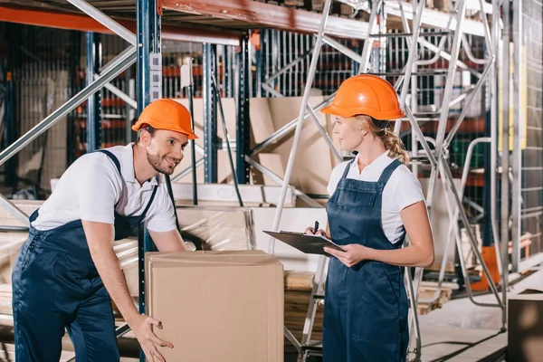 Smiling loader holding cardboard box and looking at workwoman with clipboard — Stock Photo